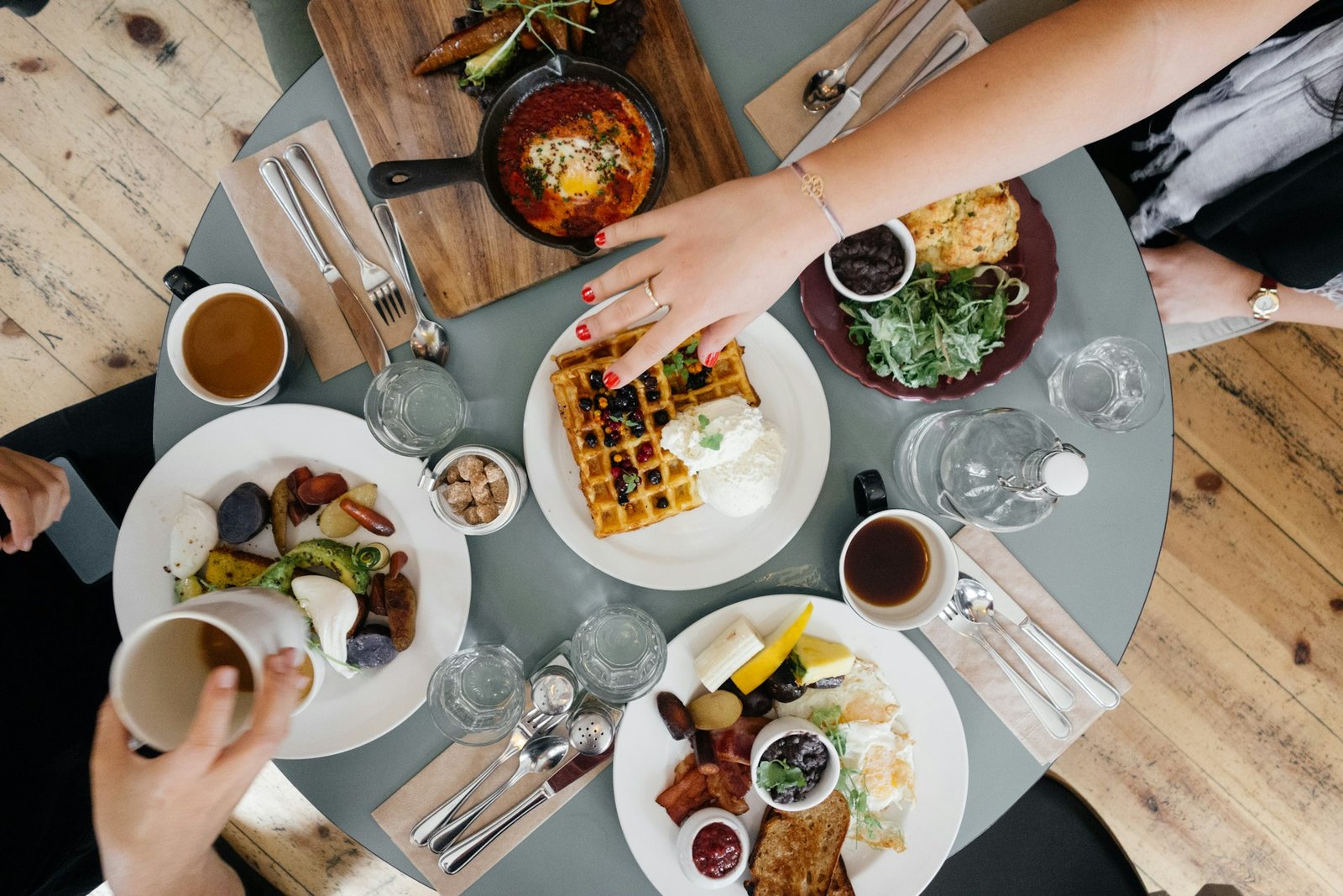 Mesa compartida con platos de desayuno y manos de varios comensales, vista desde arriba